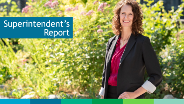 Superintendent's Report Woman in black blazer and magenta shirt smiling and standing in front of green bush with pink flowers with sunshine beaming from behind.