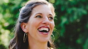 Victoria Maxwell Smiling woman with long brown hair in yellow sleeveless dress holds a microphone while standing in front of green bushes and green grass