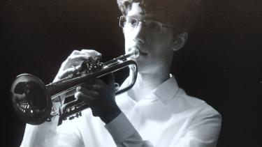 Black and white photo in partial silhouette, of young man with brown curly hair wearing white, button-up shirtplaying trumpet