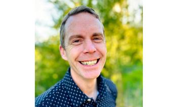 A man with short, sandy blond hair and a broad smile stands in front of some green bushes
