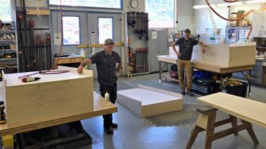 Two carpenters smile beside wooden structures in they have built in their shop