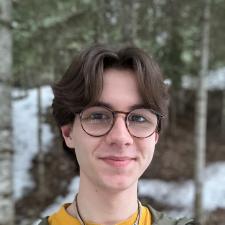 A teenaged boy with brown hair wearing glasses and a green jacket stands in front of some trees.