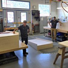 Two carpenters smile beside wooden structures in they have built in their shop